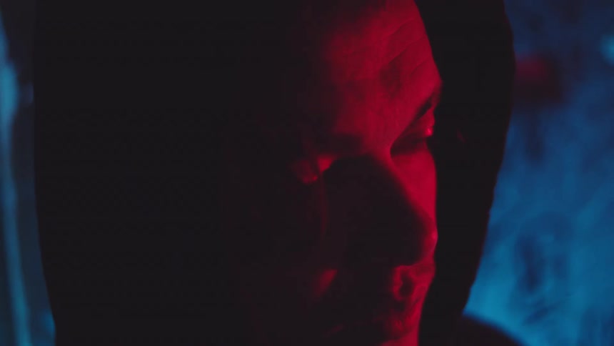 Red and blue lighting shine onto the face of a man standing while wearing a hoodie