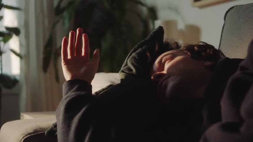 A man wearing a dark hoodie is lying on a couch resting with his hand slightly raised towards the soft afternoon sunlight illuminating his face and the living room