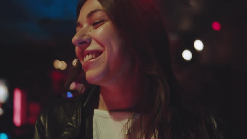 A smiling woman in a leather jacket is in a bar or restaurant illuminated by colorful background lights