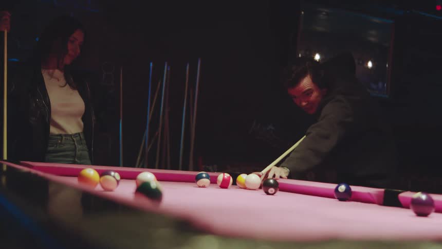 Pair playing pool on pink table with scattered balls and visible cues in dimly lit setting