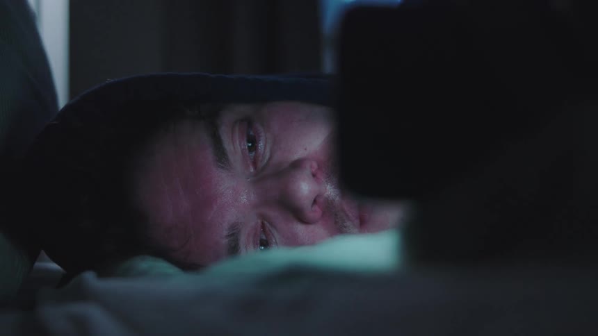 A young man wearing a hoodie lies with his head on a pillow his face showing a neutral expression as he looks to a smartphone screen before turning his head