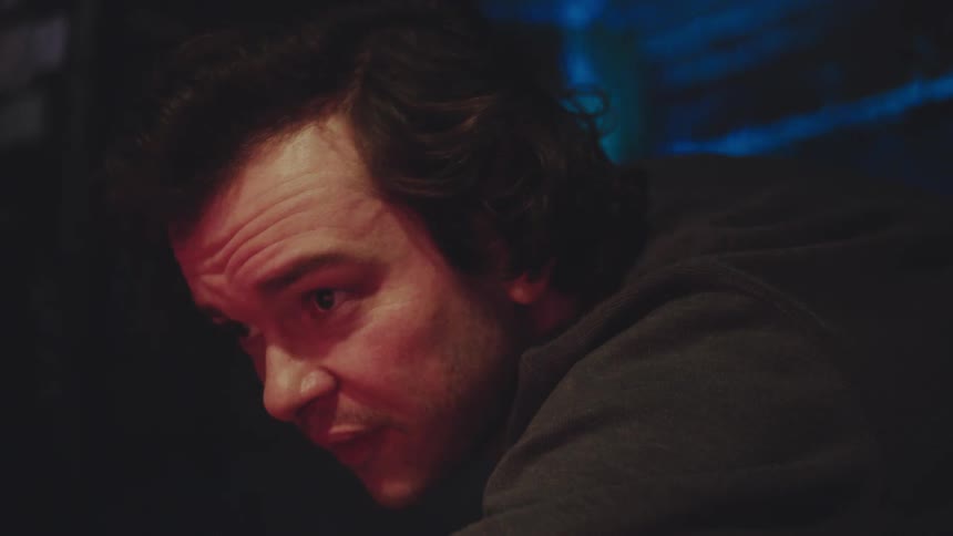A closeup shot of a mans face illuminated by red and blue light in a dark room as he looks intently and then smiles while holding a cue stick playing a game of billiards