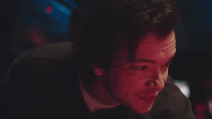 A young man leans forward his face illuminated by a striking red glow flashing a brief engaged grin at someone offcamera before slowly pulling back and looking up within a dark lively interior space