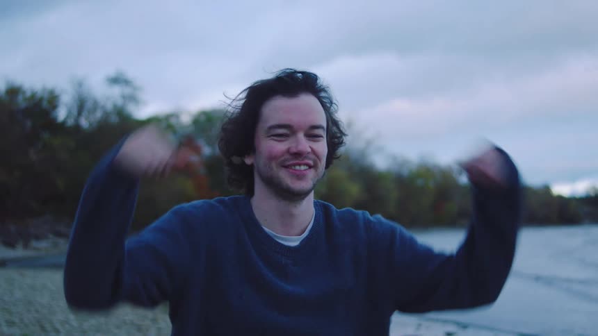 Man with dark hair in blue sweater holding silver device near lake and trees