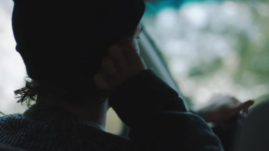 A closeup shot focuses on the back of a man wearing a black beanie and a patterned sweater while driving in a sunlit car he briefly turns his head to the side smiles and adjusts his earring or earpiece before looking back towards the road