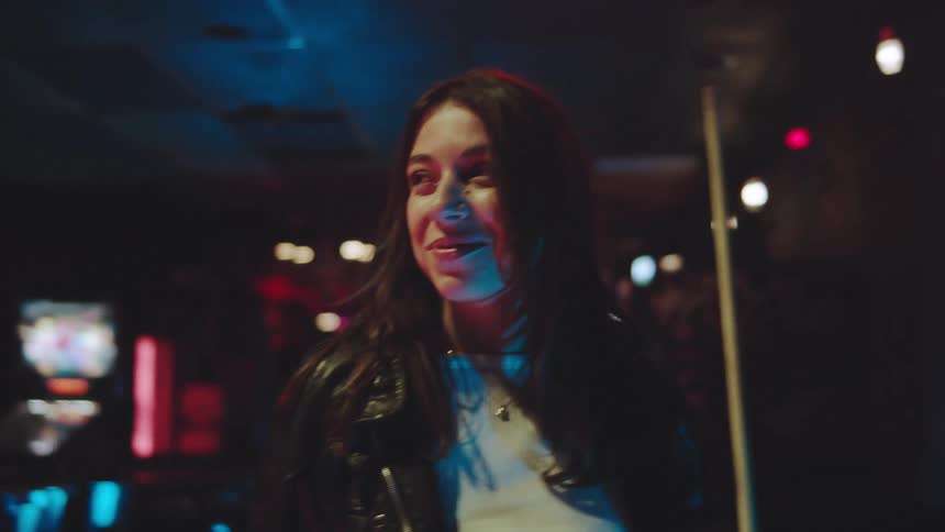 A woman with long dark hair wearing a black leather jacket smiles while socializing in a darkly lit club environment with neon accents holding a pool cue stick and appearing to talk to someone offcamera