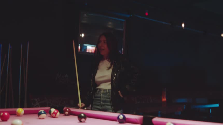 A man and a woman in a dimly lit room with blue and red neon accents play a game of pool on a bright pink table taking turns aiming and striking the cue ball