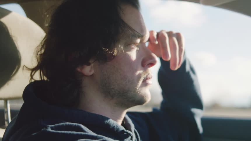 A young man drives his car in the countryside at midday with one hand on the steering wheel and the other resting on the side of his face