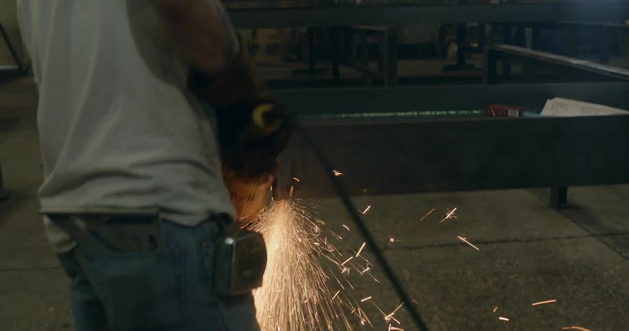 A man grinds down a metal beam in a factory as sparks fly all around