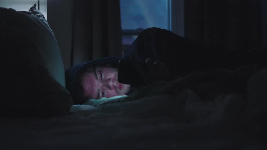 A hooded man lies in bed illuminated by his phone screen then lowers it to stare into the darkness