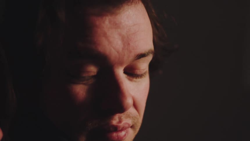 This closeup video shows the profile of a man with light stubble in dim dramatic lightinghe looks up and to the right then briefly closes his eyes and smiles before opening them again suggesting a moment of reflection or reaction