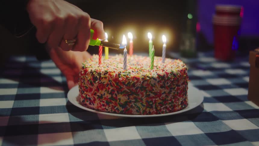 A closeup shot focuses on a persons hand using a lighter to light the small candles placed in a round birthday cake covered in colorful sprinkles on a checkered tablecloth