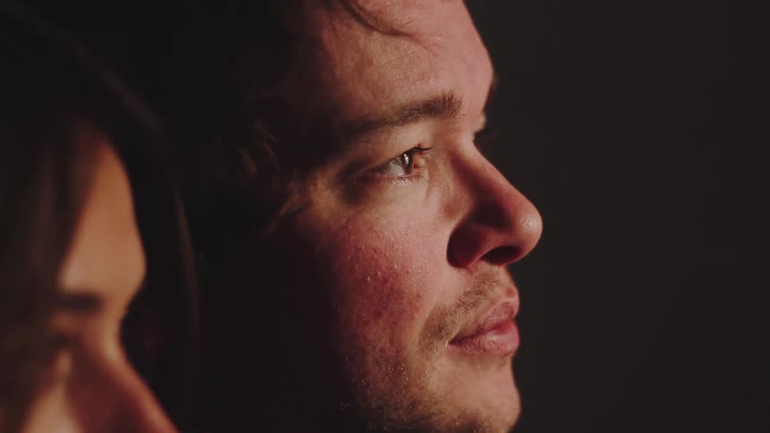 Side profile of a man lit by soft light in a dark setting with another person out of focus in the background