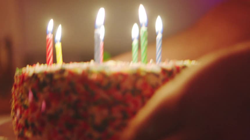 A person holds a birthday cake covered in colorful sprinkles captured in a closeup with a shallow depth of field showcasing seven brightly burning candles against a backdrop of dim colorful light