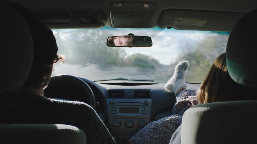 A man driving a car while his female passenger relaxes with her feet wearing fuzzy socks propped up on the dashboard during a bright casual road trip through the countryside