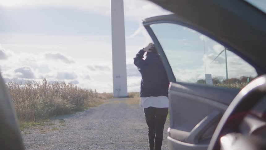 A man in a dark hoodie initially silhouetted in a car and rubbing his face steps out onto a gravel road next to a wind turbine and a field of tall grass on a bright sunny day