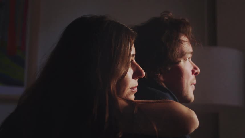 A woman approaches a man sitting alone in a dimly lit room and hugs him from behind they both look forward illuminated by a soft light from the side with the mans expression appearing sad or contemplative and the woman providing comfort