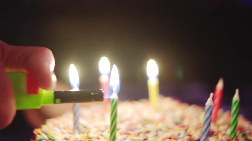 A hand lights candles on a decorated birthday cake in low light