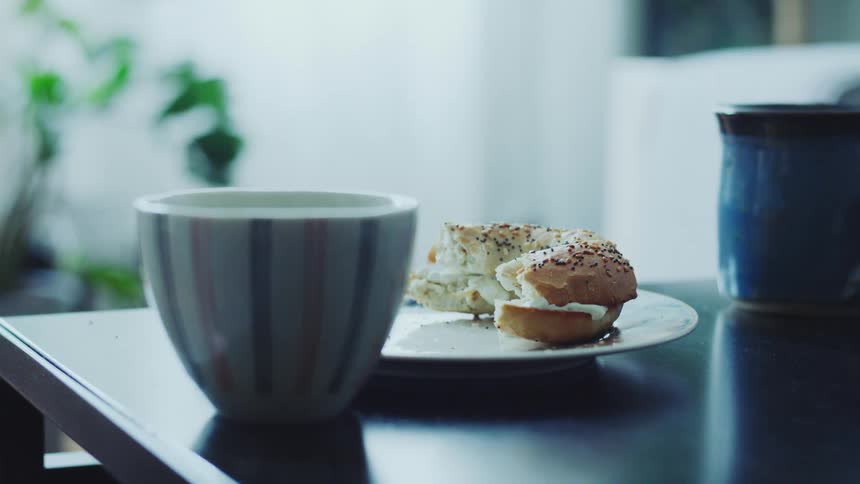 A half eaten bagel with cream cheese and poppy seeds sits on a white plate next to two coffee mugs on a dark table in soft daylight