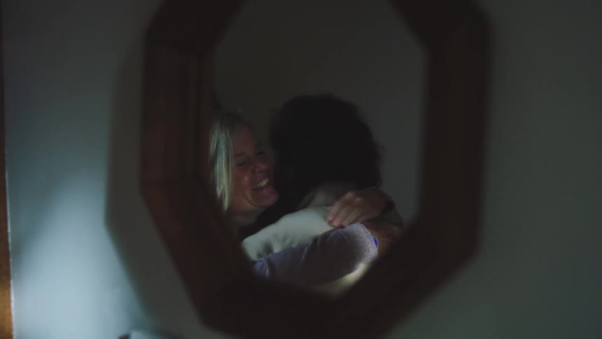 Reflected in an octagonal wall mirror a smiling woman embraces a man
