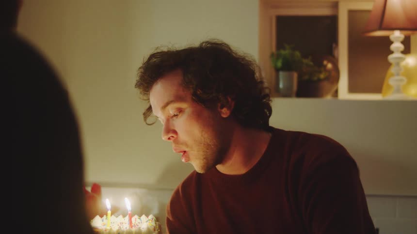 Man blowing out candles on a birthday cake at a table in an indoor setting