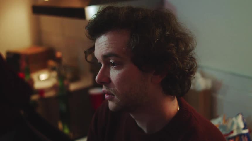 The video is a closeup sideprofile shot of a young man with dark curly hair and a reddishbrown sweater he appears to be focused on something offcamera in a dimly lit casual setting that resembles a kitchen or common area