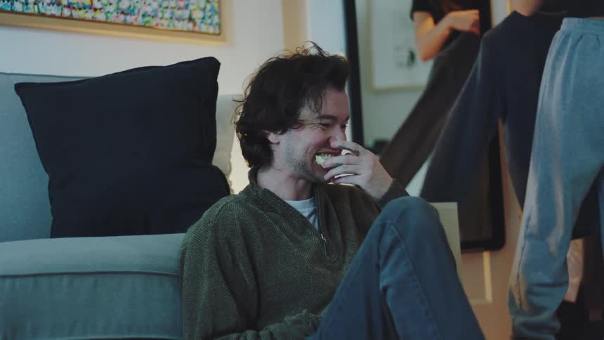 A man with dark hair sits casually on the living room floor leaning on a gray sofa eating a snack and smiling as a woman gets dressed in the background