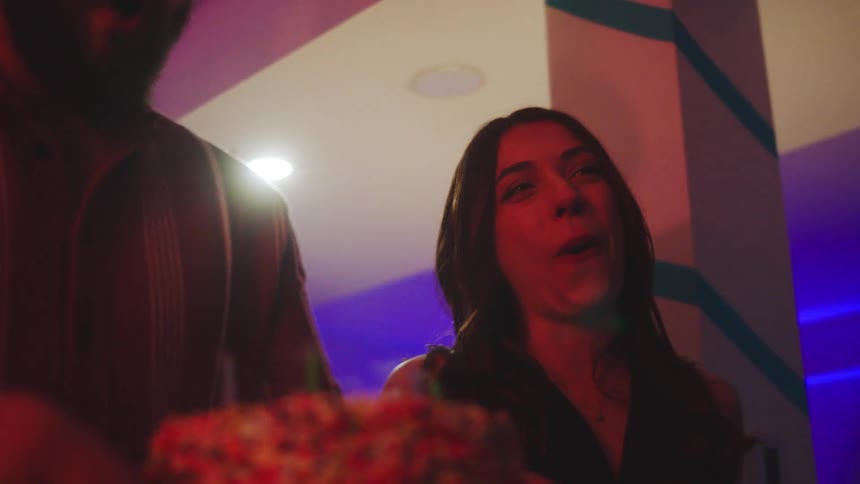 Woman smiling and clapping in celebration in front of a cake with lit candles in an indoor setting with colored lights