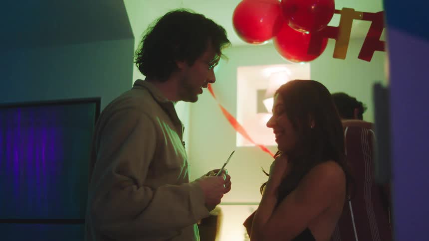 In a dimly lit house party with red balloons and a birthday banner a man in a tan shirt talks to a smiling woman in a black top sharing a brief intimate moment before they turn and join the celebration