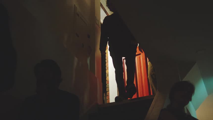 A young man climbs a staircase and opens the door to let another young man inside they bump fists then walk back down the stairs into a room filled with people
