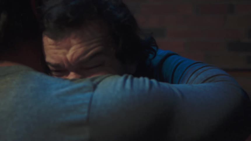 A man with a serious expression stands against a brick wall then steps forward to embrace another person he tightly hugs the other person burying his face in their shoulder in a moment of emotional support