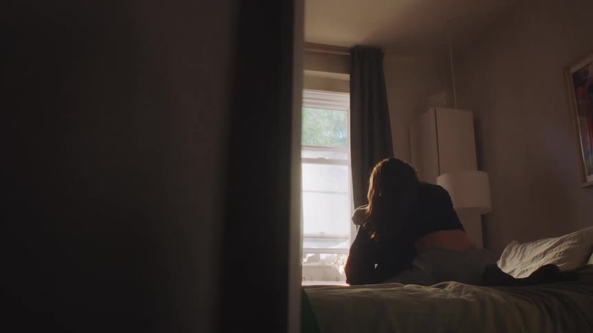 A young woman walks and sits behind a man hugging him on the bed as the sunrise brightens the room