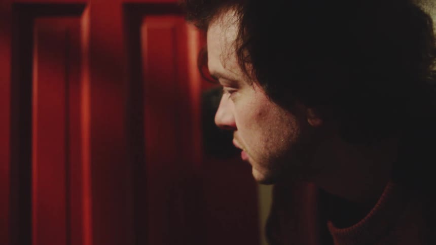 A man in a dark red shirt is first seen quickly tying the laces of his hightop sneaker on a patterned rug then lifts his head into a dramatic closeup beside a bright red door