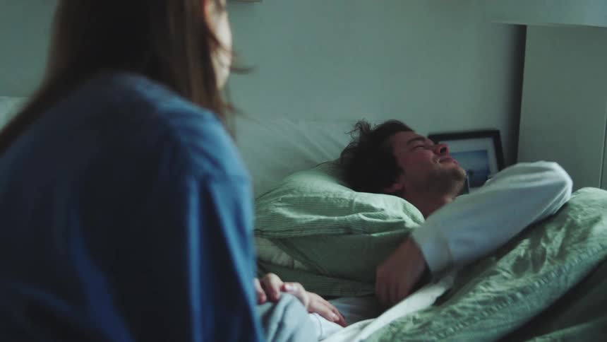 Young adult woman helping a young adult man who is struggling to wake up in a bedroom indoors during day time