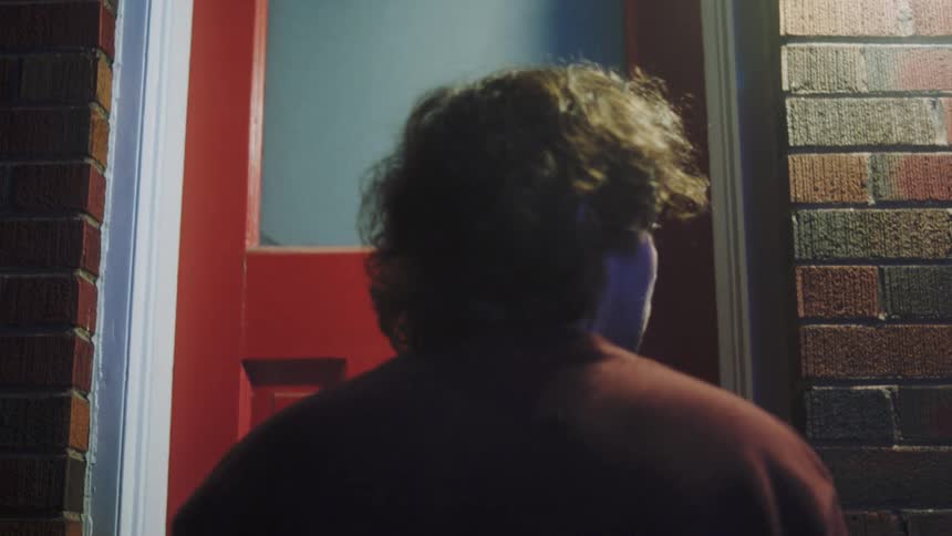 A young man stands outdoors at night knocking on a red door and a man opens it as they hug each other