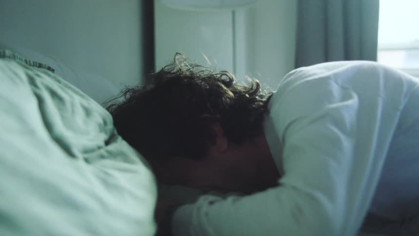 The video shows a person sleeping under covers another person enters the frame leans in and gently touches the first persons back who then stirs and begins to wake up