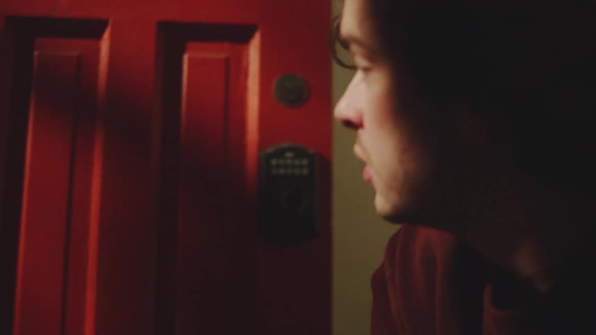 A young man puts on sneakers while sitting beside a wooden door lost in thought