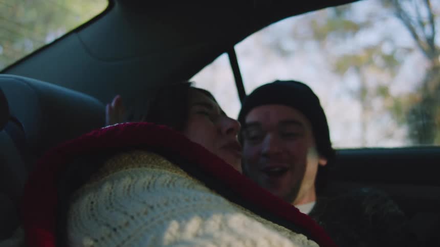 A man and woman wrapped in a white and red blanket are cuddling and smiling in the backseat of a moving car framed by the bright outoffocus outdoor scenery