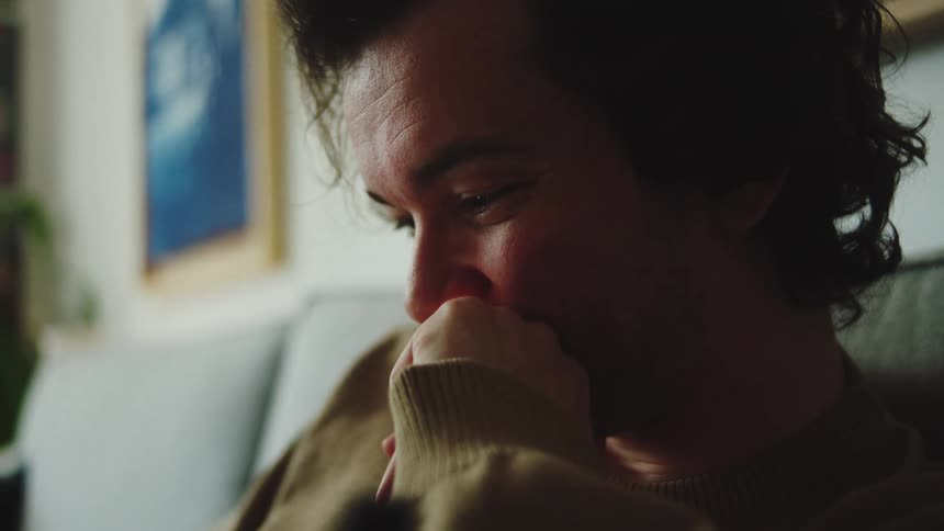 A young man sits on a couch indoors looking down at his phone and smiling as he brings his hand to his mouth