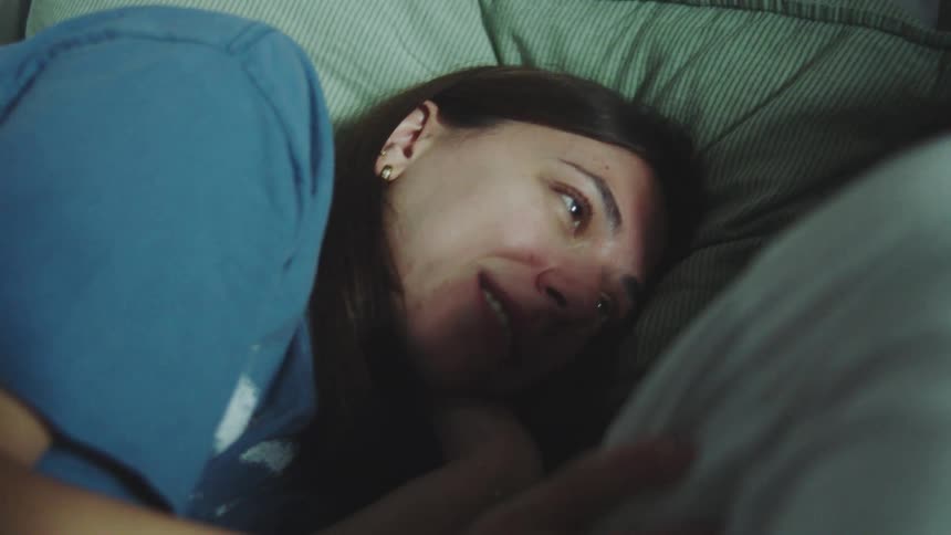 A close view shows a woman in a blue shirt smiling and talking gently to a man lying next to her on green pillows in a dimly lit bedroom