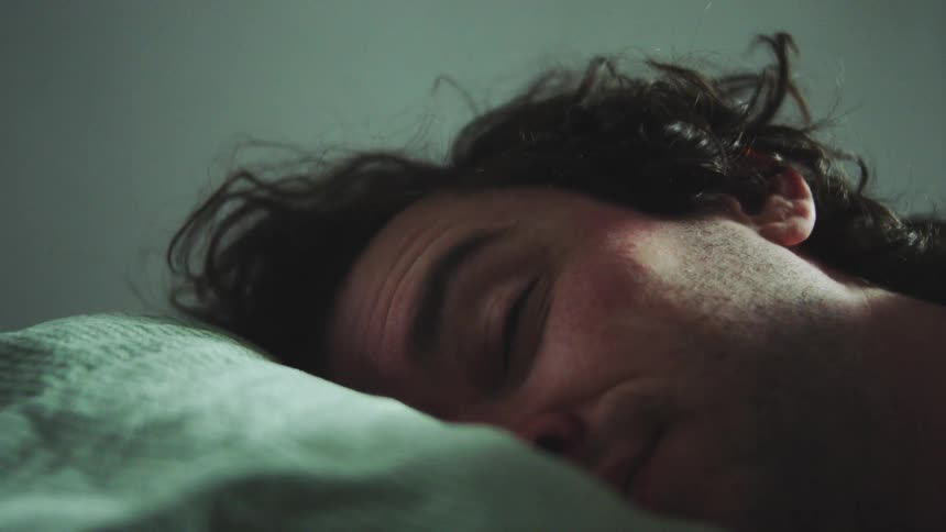 A close view of a man with dark curly hair sleeping soundly on a pillow with his eyes closed in a dimly lit dark green room