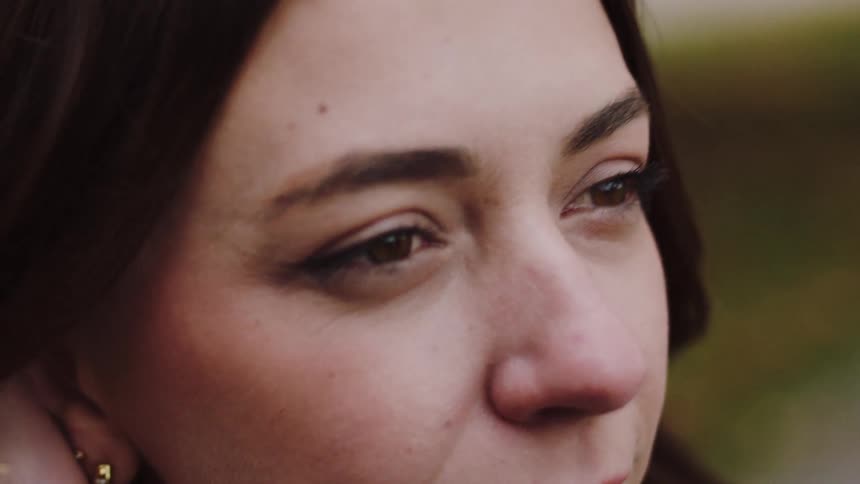 A woman with dark hair and noticeable eyeliner is shown from a slight profile angle looking off into the distancethe shot is relatively tight on her face and is outdoors with soft lighting and a blurred background