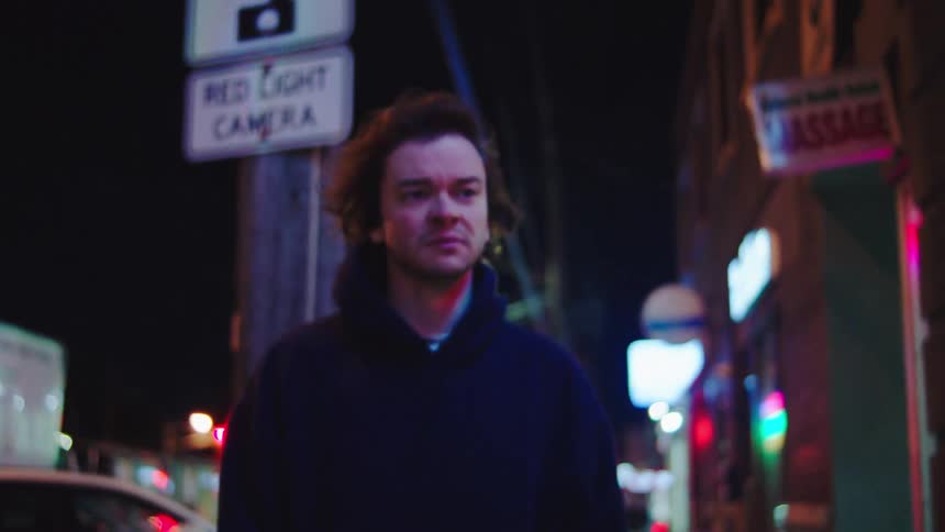 A young man walks along the sidewalk at night wearing a black jacket