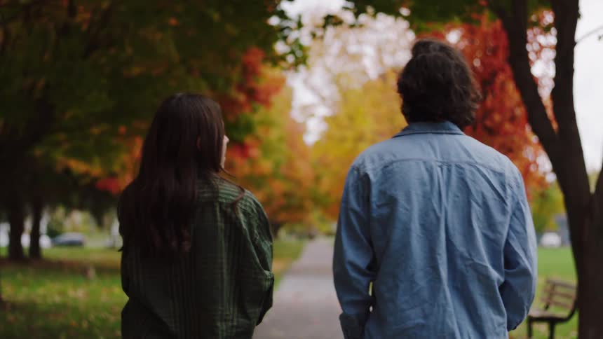 Young adult man and young adult woman walking next to each other while talking on a park outdoors during day time