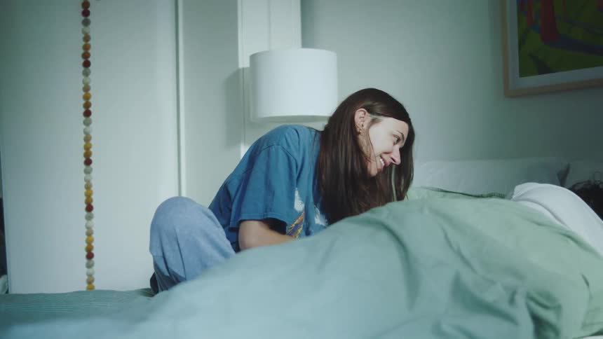 Couple sitting on a bed with a woman smiling while looking at the person lying beside her