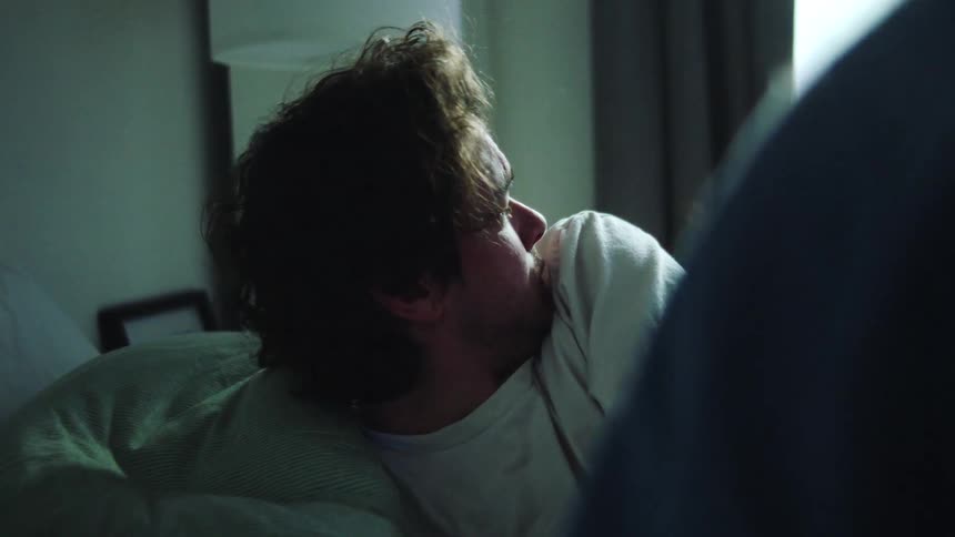 A dark interior shot shows a man with messy hair sleeping soundly in a white shirt on a striped pillow as a person gently wakes him with a touch on the arm