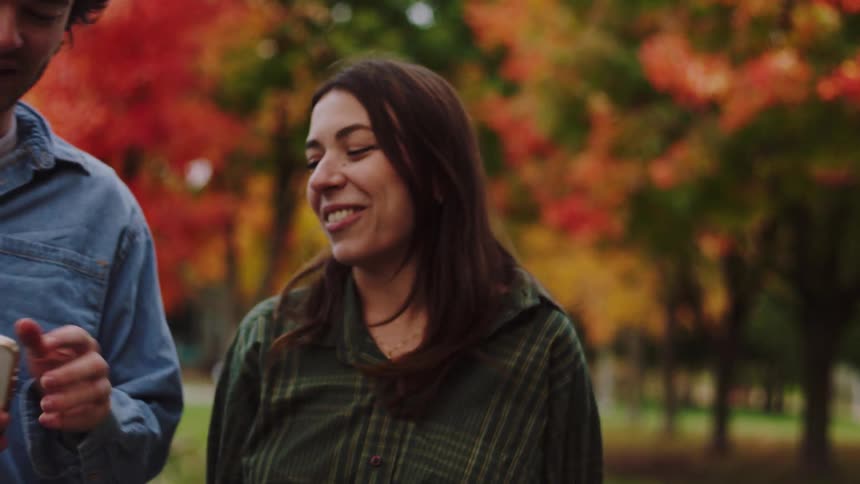A young woman with long dark hair is seen laughing and talking animatedly with a young man in a closeup shot as they walk through a park with the background showcasing bright red and orange autumn foliage