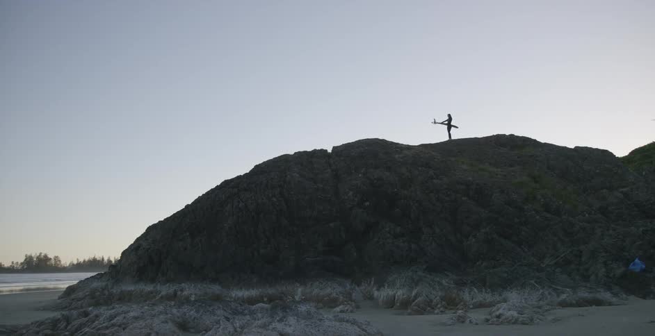 A person holding a surfboard stands on top of a mountain by the ocean