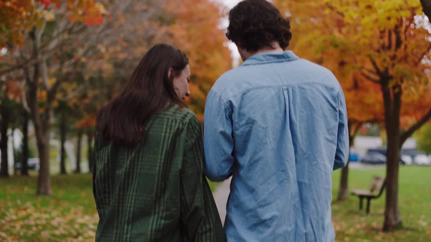 A couple walk in a public park with trees lining the pathway and fallen leaves from autumn at midday