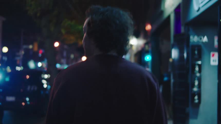 The camera tracks closely behind a person with dark curly hair wearing a deep red or maroon sweatshirt as they walk down a city street at night bright streetlights and the blurred colorful bokeh of car headlights and storefronts illuminate the scene